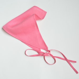 Puede incluir: Un pañuelo rosa brillante, de forma triangular, con lazos largos y finos. La tela parece ligera y los bordes están cuidadosamente acabados. El pañuelo está doblado y atado, listo para usar. Un accesorio versátil.