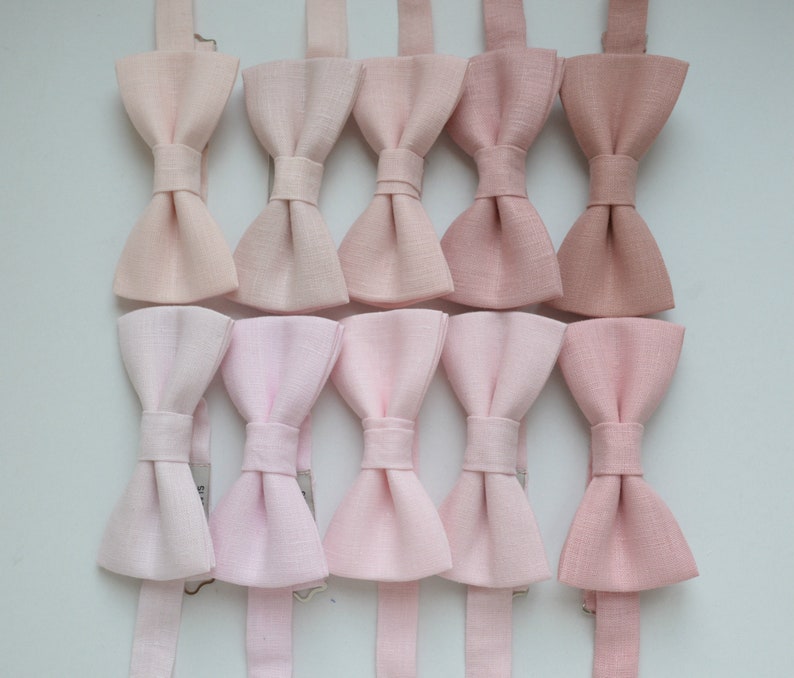 May include: A collection of ten bow ties in various shades of pink, from light blush to deeper rose. Each bow tie is neatly arranged, showcasing the different hues and textures of the fabric. The ties are designed for formal wear.