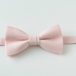 May include: A light pink bow tie made of textured fabric. The bow tie is centered and has a classic shape with a pre-tied knot. The adjustable neckband is visible, with a metal clasp. The background is plain white.