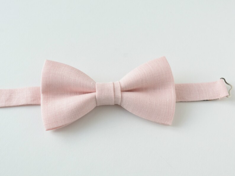 May include: A light pink bow tie made of textured fabric. The bow tie is centered and has a classic shape with a pre-tied knot. The adjustable neckband is visible, with a metal clasp. The background is plain white.
