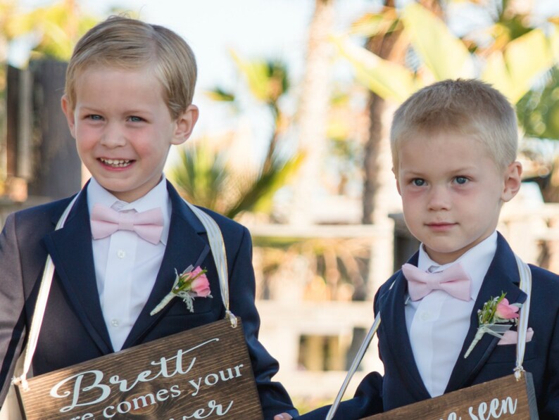 May include: Two young boys in navy blue suits with pink bow ties and boutonnieres. Each boy holds a wooden sign with white text. The sign on the left reads "Brett here comes your..." and the sign on the right reads "...seen".