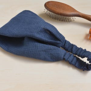 May include: A navy blue fabric headband with an elastic band. The headband has a textured pattern and is lying on a light-colored surface. A wooden hairbrush and a brown hair clip are also visible.