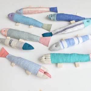 Könnte beinhalten: Eine Sammlung handgefertigter Stofffische in verschiedenen Farben und Mustern. Die Fische haben blaue, rosa und grüne Schwänze mit Körpern aus gestreiften und unifarbenen Stoffen. Jeder Fisch hat kleine, beige Stoffflossen.