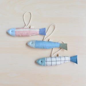 Könnte beinhalten: Drei Stofffisch-Ornamente mit blauen, roten und weißen Mustern. Jeder Fisch hat einen hellblauen Kopf und eine Schwanzflosse, mit einer Schlaufe aus naturfarbenem Bindfaden zum Aufhängen. Auf einer hellen Holzoberfläche angeordnet.