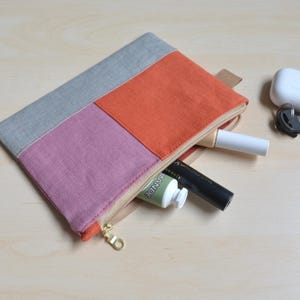 May include: A rectangular zippered pouch featuring a color-blocked design with a gray top section, a mauve middle section, and an orange bottom section. The pouch is partially open, revealing a white tube, a black tube, and a small green tube inside.
