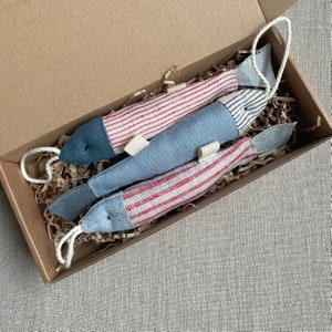 Puede incluir: Tres juguetes de tela con forma de pez en una caja de cartón. Dos peces tienen cuerpos a rayas rojas y blancas, mientras que el otro es azul liso. Todos tienen aletas grises y están rellenos con un lazo de cordón blanco para colgar. La caja está llena de papel triturado.