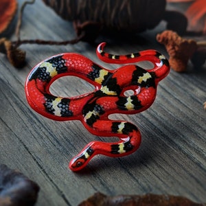 May include: A red and black snake figurine with a detailed painted design. The snake is curled in a circle with its head facing the right. The figurine is made of a smooth, glossy material.