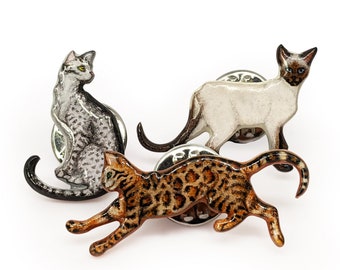 Könnte beinhalten: Drei Katzen-Anstecknadeln aus Emaille. Eine zeigt eine graue Tabby-Katze im Sitzen, eine eine weiße Siamkatze beim Gehen und eine eine braune Tabby-Katze beim Laufen.