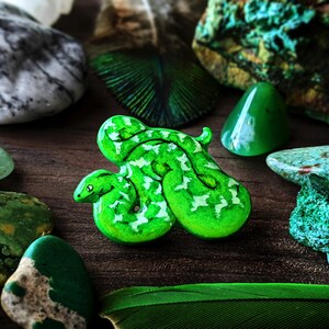 A bright green emerald tree boa pin surrounded by blue-green stones, and deep green feathers on a wooden surface.