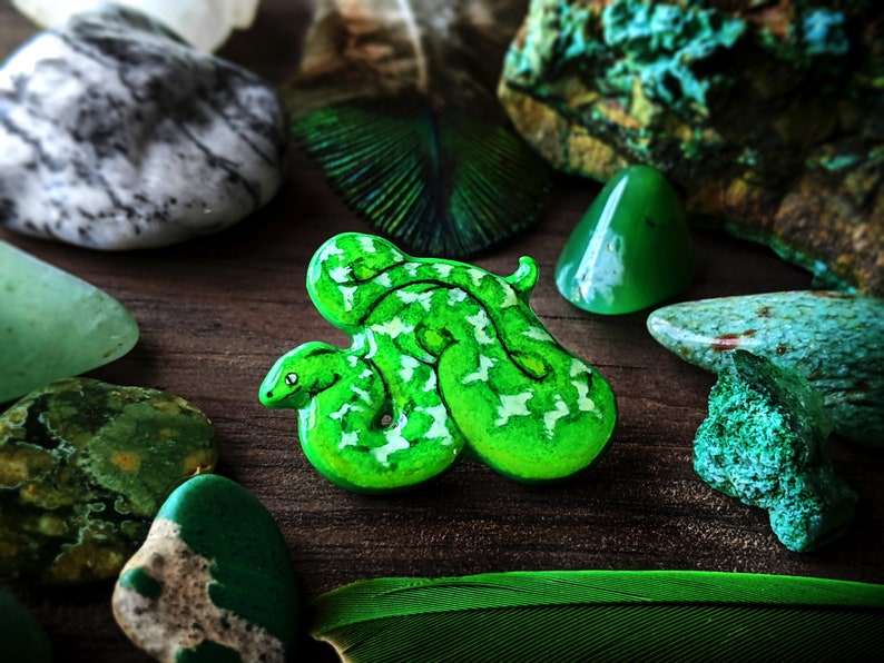 A bright green emerald tree boa pin surrounded by blue-green stones, and deep green feathers on a wooden surface.