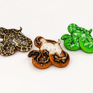Three snake pins - a Burmese python, pied ball python and emerald tree boa on a white surface.