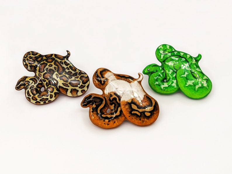 Three snake pins - a Burmese python, pied ball python and emerald tree boa on a white surface.