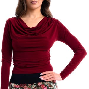 Op de afbeelding: Een bordeauxrode top met lange mouwen en een col. De top wordt gecombineerd met een broek met bloemenprint en een zwarte tailleband. De outfit staat tegen een witte achtergrond.