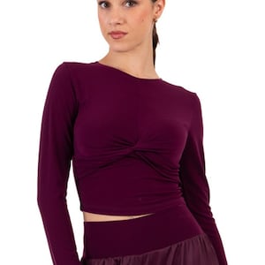 Puede incluir: Una blusa de manga larga de color burdeos con un detalle de nudo en la parte delantera. La blusa es corta y tiene una silueta ajustada.