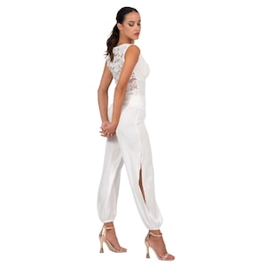 May include: A white jumpsuit featuring a sleeveless bodice with a lace back panel and long, loose-fitting trousers with high side slits at the legs. The garment has a cinched waist and gathered cuffs at the ankles, paired with metallic gold high-heeled sandals.