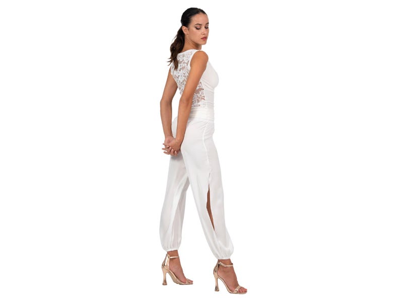 May include: A white jumpsuit featuring a sleeveless bodice with a lace back panel and long, loose-fitting trousers with high side slits at the legs. The garment has a cinched waist and gathered cuffs at the ankles, paired with metallic gold high-heeled sandals.