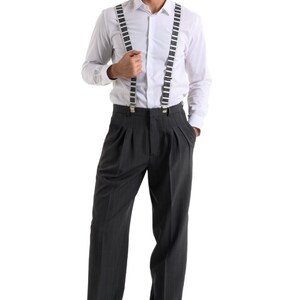May include: A man wearing a white long-sleeved shirt with black and white piano key suspenders and gray pinstripe pants. He is wearing black shoes with white accents.