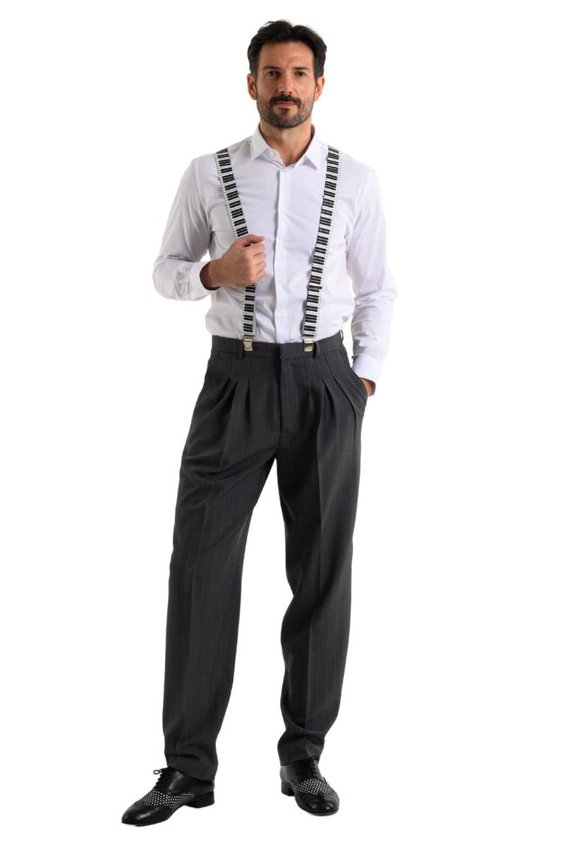 May include: A man wearing a white long-sleeved shirt with black and white piano key suspenders and gray pinstripe pants. He is wearing black shoes with white accents.