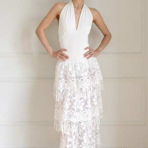 May include: A white halter top with a deep V-neckline paired with a white layered maxi skirt featuring delicate lace and fringe details.