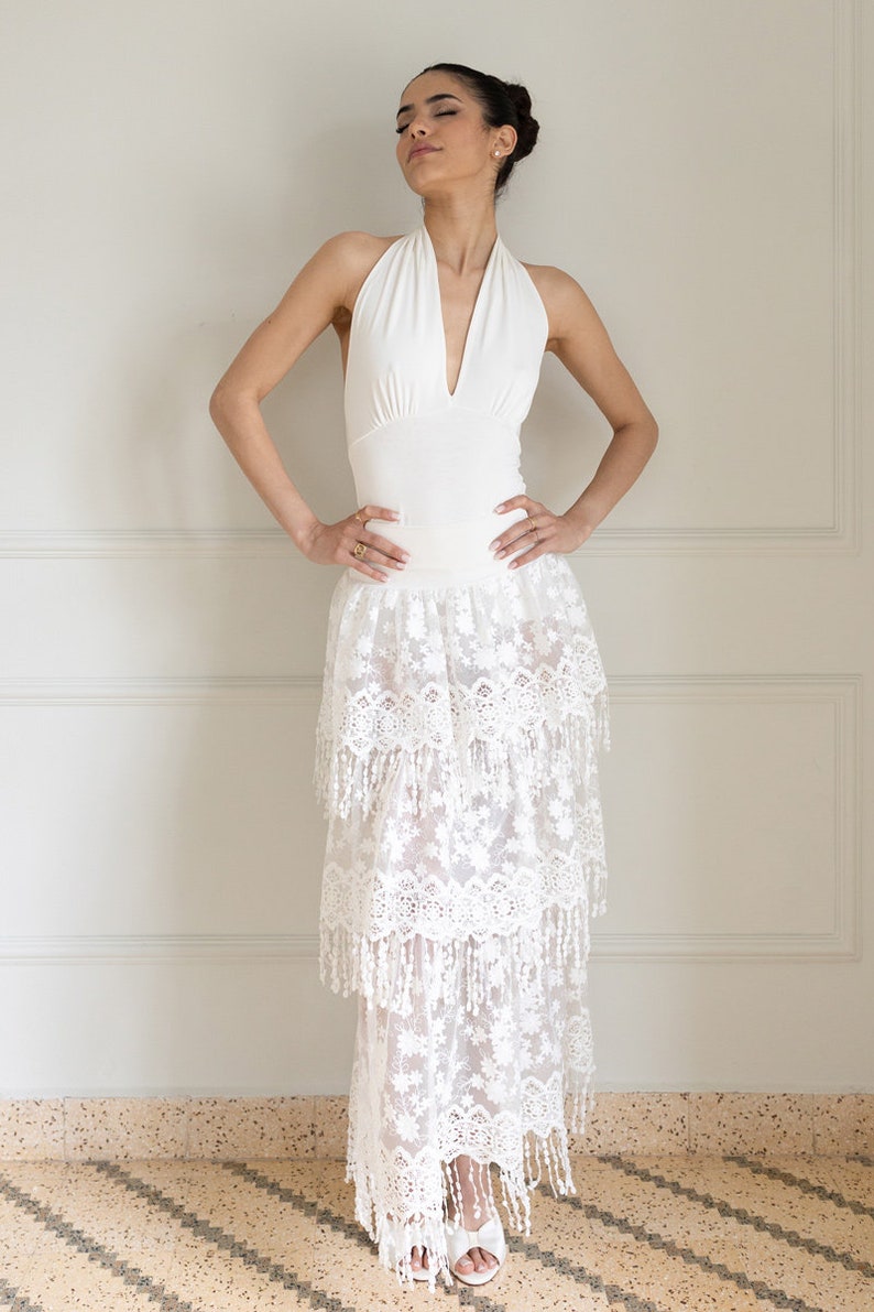 May include: A white halter top with a deep V-neckline paired with a white layered maxi skirt featuring delicate lace and fringe details.