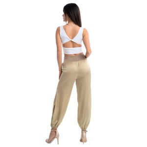 May include: A white sleeveless crop top with a V-neckline and a twisted back detail paired with high-waisted beige trousers featuring a wide elastic waistband, a relaxed fit, side leg slits, and gathered elastic cuffs at the ankles.