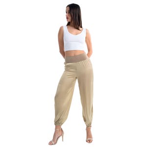 May include: A white sleeveless crop top with a scoop neckline paired with high-waisted beige satin-finish joggers featuring a wide elasticized waistband and gathered elastic cuffs at the ankles, worn with beige heeled sandals.
