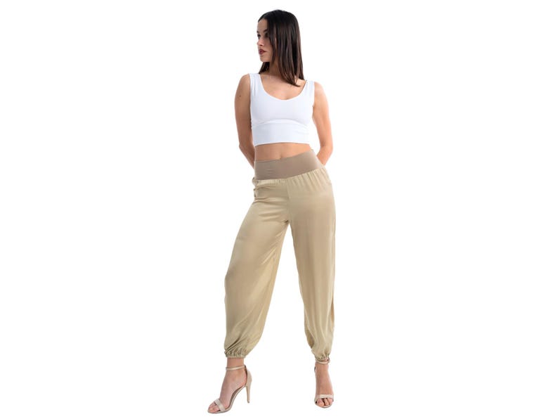 May include: A white sleeveless crop top with a scoop neckline paired with high-waisted beige satin-finish joggers featuring a wide elasticized waistband and gathered elastic cuffs at the ankles, worn with beige heeled sandals.