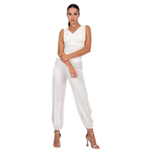 May include: A white sleeveless jumpsuit featuring a cowl neckline and a fitted bodice that transitions into loose-fitting, tapered trousers with gathered elasticized cuffs at the ankles. The garment is made of a smooth, solid white fabric.