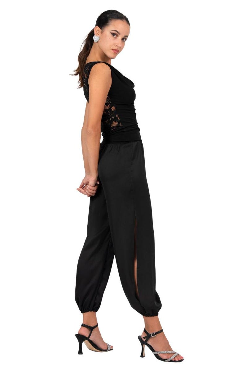 May include: A black sleeveless top featuring a cowl neckline and intricate lace side panels, paired with matching black loose-fitting trousers that have side slits and elasticized cuffs at the ankles, worn with black strappy high-heeled sandals.