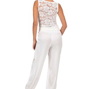 May include: A white sleeveless jumpsuit featuring a lace bodice with a floral pattern and a solid white lower section. The pants have a wide waistband and gathered elastic cuffs at the ankles, paired with metallic gold high-heeled sandals.