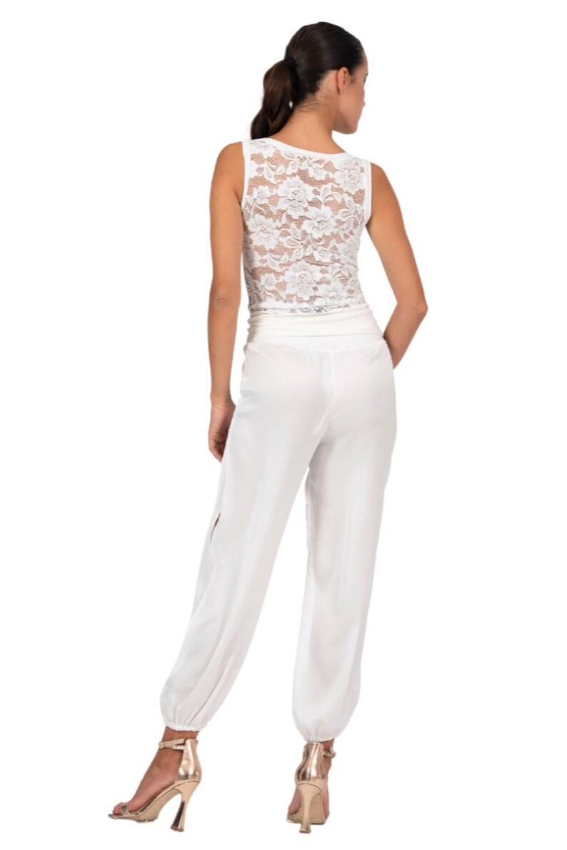 May include: A white sleeveless jumpsuit featuring a lace bodice with a floral pattern and a solid white lower section. The pants have a wide waistband and gathered elastic cuffs at the ankles, paired with metallic gold high-heeled sandals.