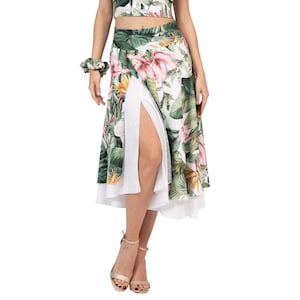 May include: A floral print skirt with a high slit and white underlay. The skirt features a tropical pattern with pink, green, and yellow flowers and leaves. A matching scrunchie is visible on the wrist. The outfit is completed with gold sandals.