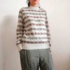 Peut inclure: Un pull crème avec des bandes horizontales de motifs rouges, verts et bleus. Le pull a un col montant et des manches longues. La personne sur l'image porte un pantalon vert olive.