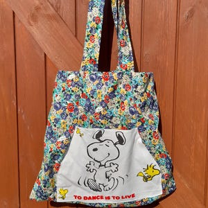 May include: A floral print tote bag with a white pocket featuring a black and white Snoopy graphic and the text "TO DANCE IS TO LIVE." The bag has matching floral handles and a blue, red, and yellow floral pattern.