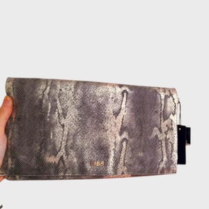 May include: A grey and gold clutch bag with a snakeskin pattern. The rectangular bag has a fold-over flap. The brand name "IBA" is visible on the front. A black tag hangs from the side.
