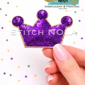 May include: A purple sequined crown patch with gold trim is held against a white background with colorful confetti. The crown has three rounded points and the text "Stitch NOLA" is visible. The patch is for embroidery and printing.