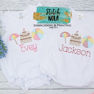 May include: Two white baby onesies with embroidered beach-themed designs. The onesies feature a sandcastle, beach umbrella, and beach ball. The names "Evey" and "Jackson" are embroidered on the onesies.