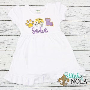 May include: White dress with a purple and gold embroidered tiger, paw print, and the state of Louisiana. The dress has a ruffle at the bottom and the name "Sadie" is embroidered on the front.