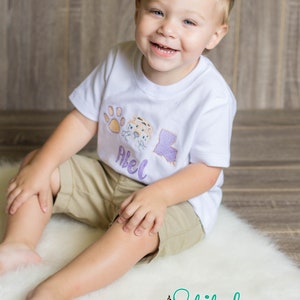 May include: A young boy wearing a white t-shirt with an embroidered paw print, tiger face, and the state of Louisiana. The shirt also has the name "Abel" embroidered on it. The boy is sitting on a white fluffy rug.