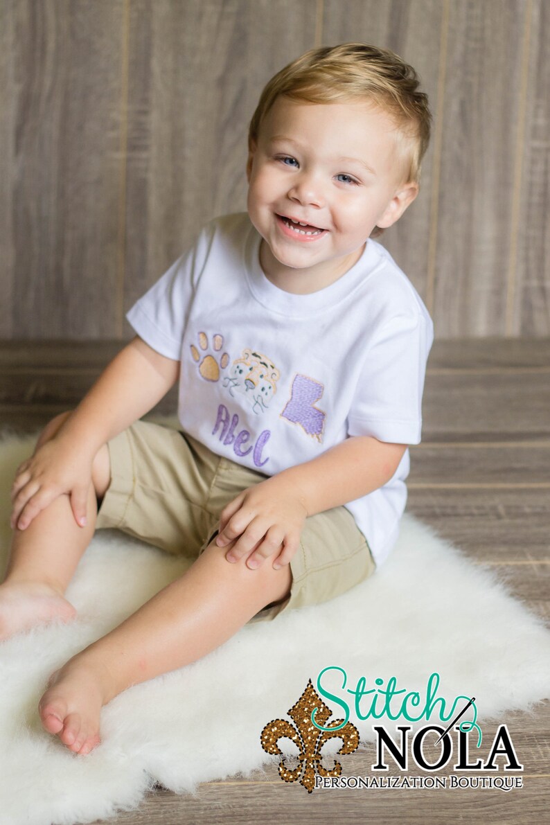 May include: A young boy wearing a white t-shirt with an embroidered paw print, tiger face, and the state of Louisiana. The shirt also has the name "Abel" embroidered on it. The boy is sitting on a white fluffy rug.