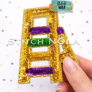 May include: A gold sequined letter "A" with purple and green accents, likely for Mardi Gras. The letter is embellished with gold sequins and features horizontal stripes of purple and green sequins. The image includes the text "Stitch NOLA".