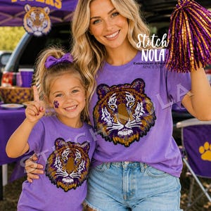 May include: Two matching purple t-shirts featuring a large sequined tiger head graphic on the front. The shirts are paired with light blue denim shorts. One shirt is sized for an adult and the other for a child. The text Stitch NOLA is visible.