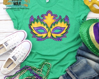 May include: Green t-shirt featuring a purple and gold Mardi Gras mask design. The shirt is laid flat on a white wooden surface with a pair of white trainers, a pair of blue jeans, and a green and gold Mardi Gras hat.