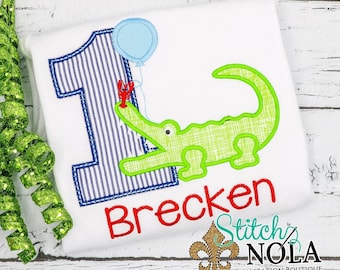 May include: A green alligator holding a red lobster with a blue balloon. The number one is in the background with blue and white stripes. The name Brecken is embroidered in red below the alligator. The text Stitch Nola Personalization Boutique is below the name.