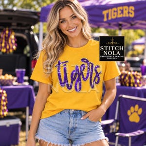 May include: A mustard yellow short-sleeved t-shirt featuring the word Tigers in purple script lettering. The lettering is embellished with purple sequins and outlined in silver. The shirt has a crew neckline and a standard fit.