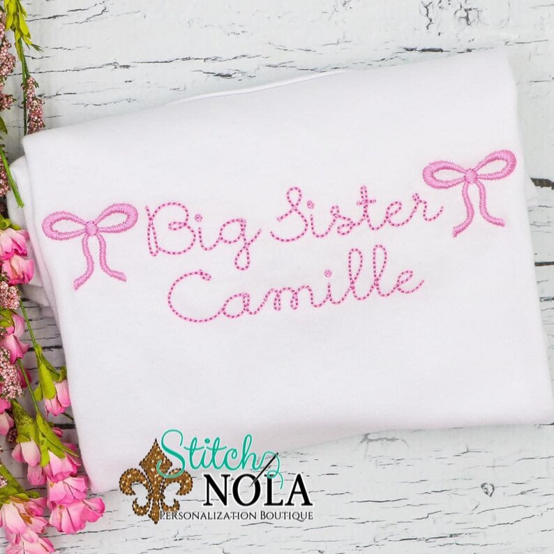 May include: White t-shirt with pink embroidered text that reads "Big Sister Camille" with two pink bows on either side of the text.