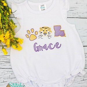 May include: White baby onesie with purple and yellow embroidery. The embroidery includes a paw print, a tiger face, the state of Louisiana, and the name "Grace".