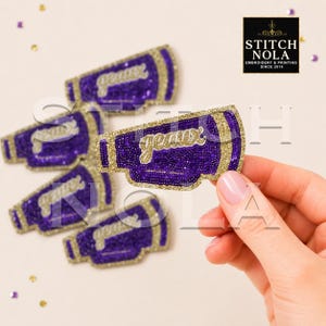 May include: A collection of purple and gold glitter megaphone-shaped patches featuring the word geaux in gold script. The patches have a textured, sparkling finish with gold borders. A logo in the corner reads STITCH NOLA EMBROIDERY & PRINTING SINCE 2014.