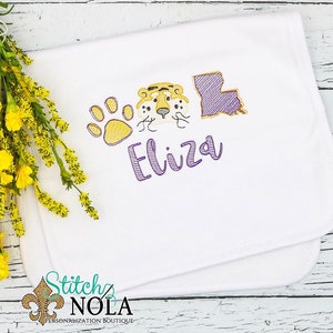 May include: White baby blanket with purple embroidery of a tiger face, paw print, and the state of Louisiana. The name "Eliza" is embroidered in purple below the images.