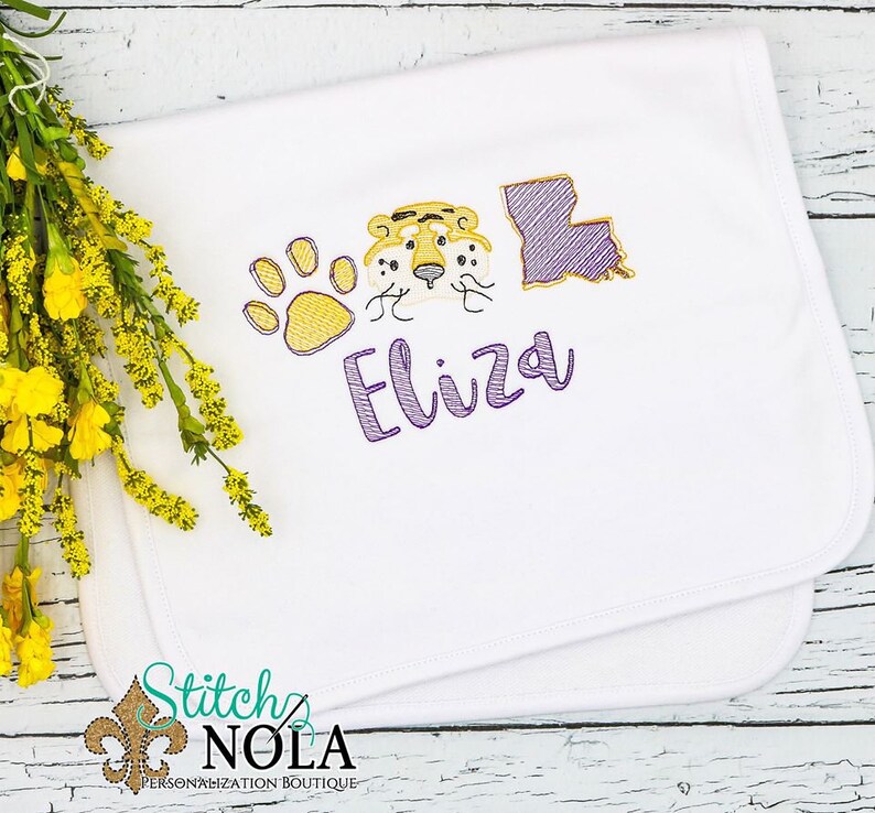 May include: White baby blanket with purple embroidery of a tiger face, paw print, and the state of Louisiana. The name "Eliza" is embroidered in purple below the images.